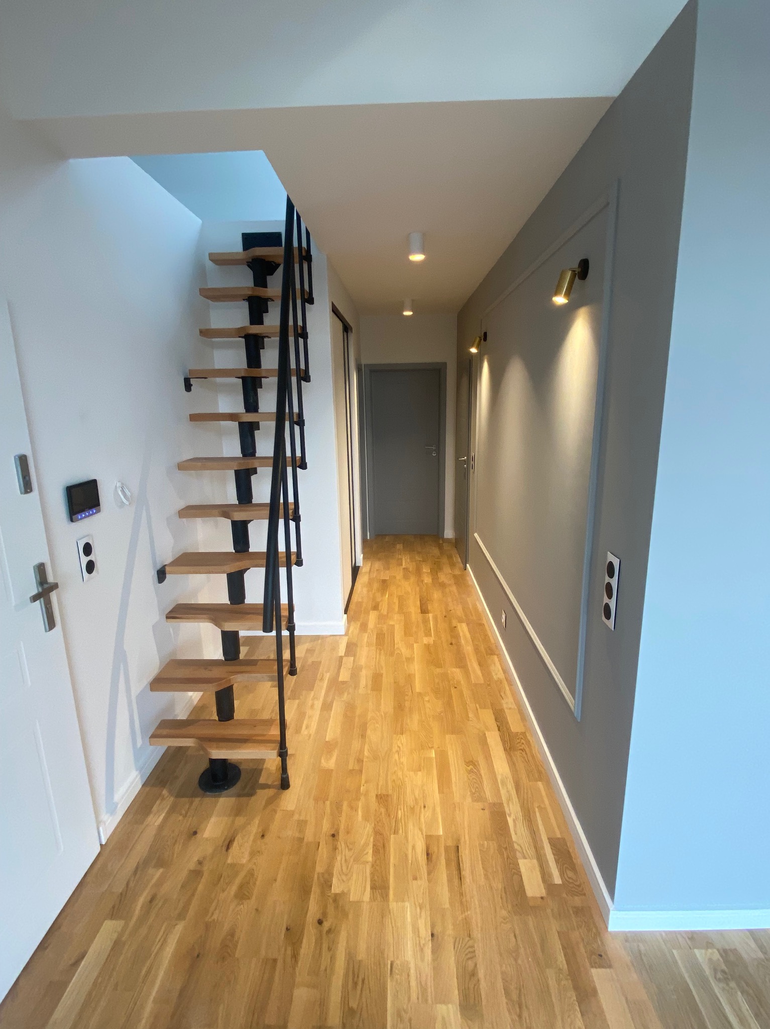 Wnętrze korytarza z drewnianą podłogą, schodami na konstrukcji z czarnego metalu, szarą ścianą z dekoracyjną listwą i złotymi lampkami punktowymi.