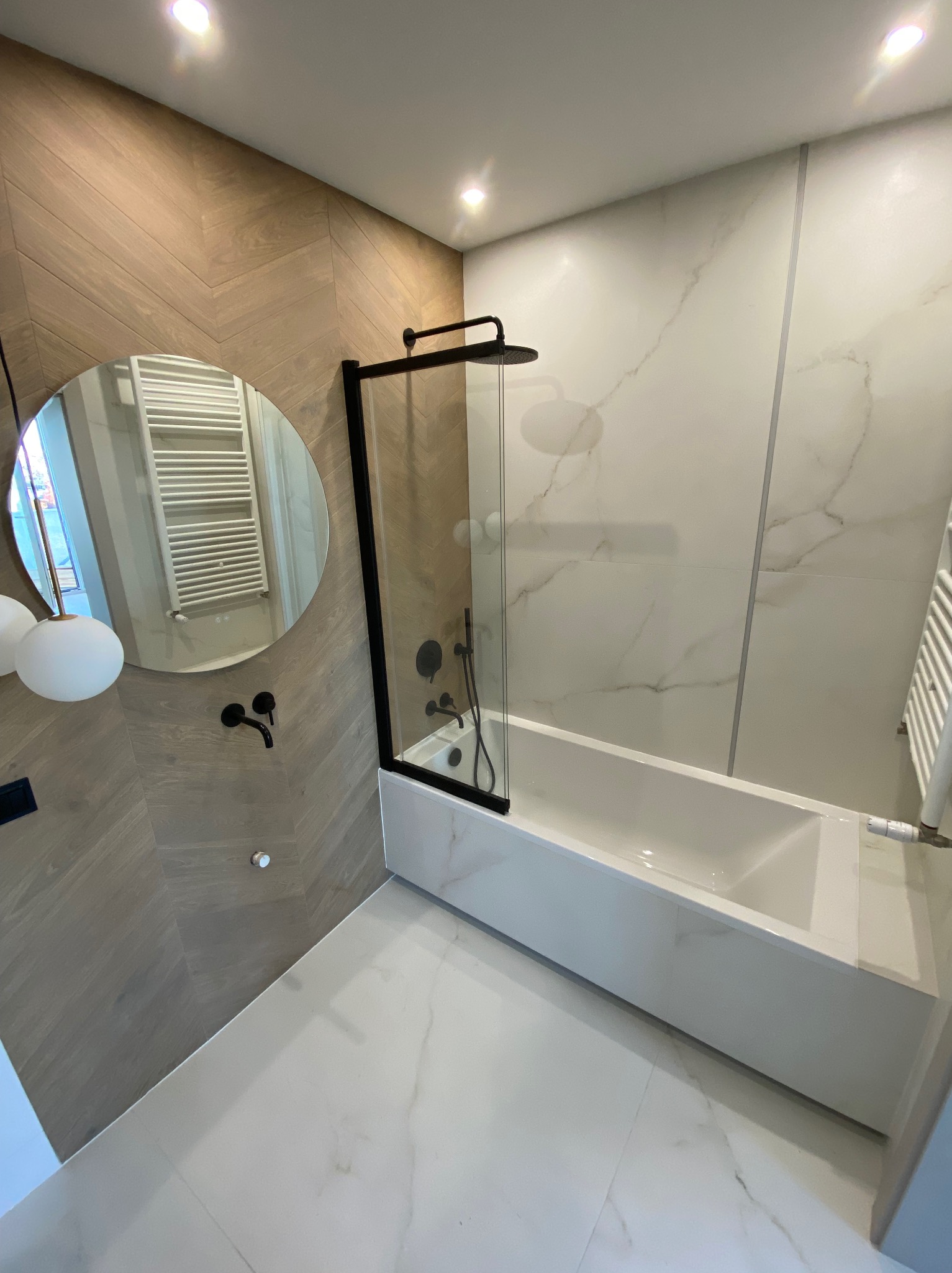 Nowoczesna łazienka z wanną za szklanym parawanem w czarnej ramie, okrągłe lustro z czarną armaturą na ścianie wykończonej drewnopodobną płytką ułożoną w jodełkę, białe płytki imitujące marmur...