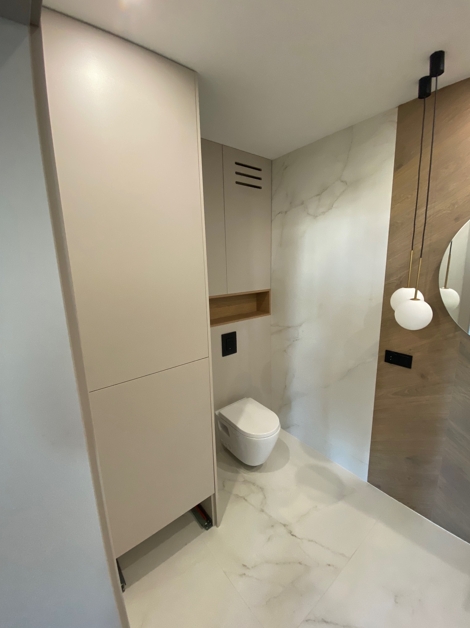 Nowoczesna łazienka z białymi marmurowymi płytkami, podwieszaną toaletą, beżowymi szafkami i okrągłym lustrem podświetlonym wiszącymi lampami.