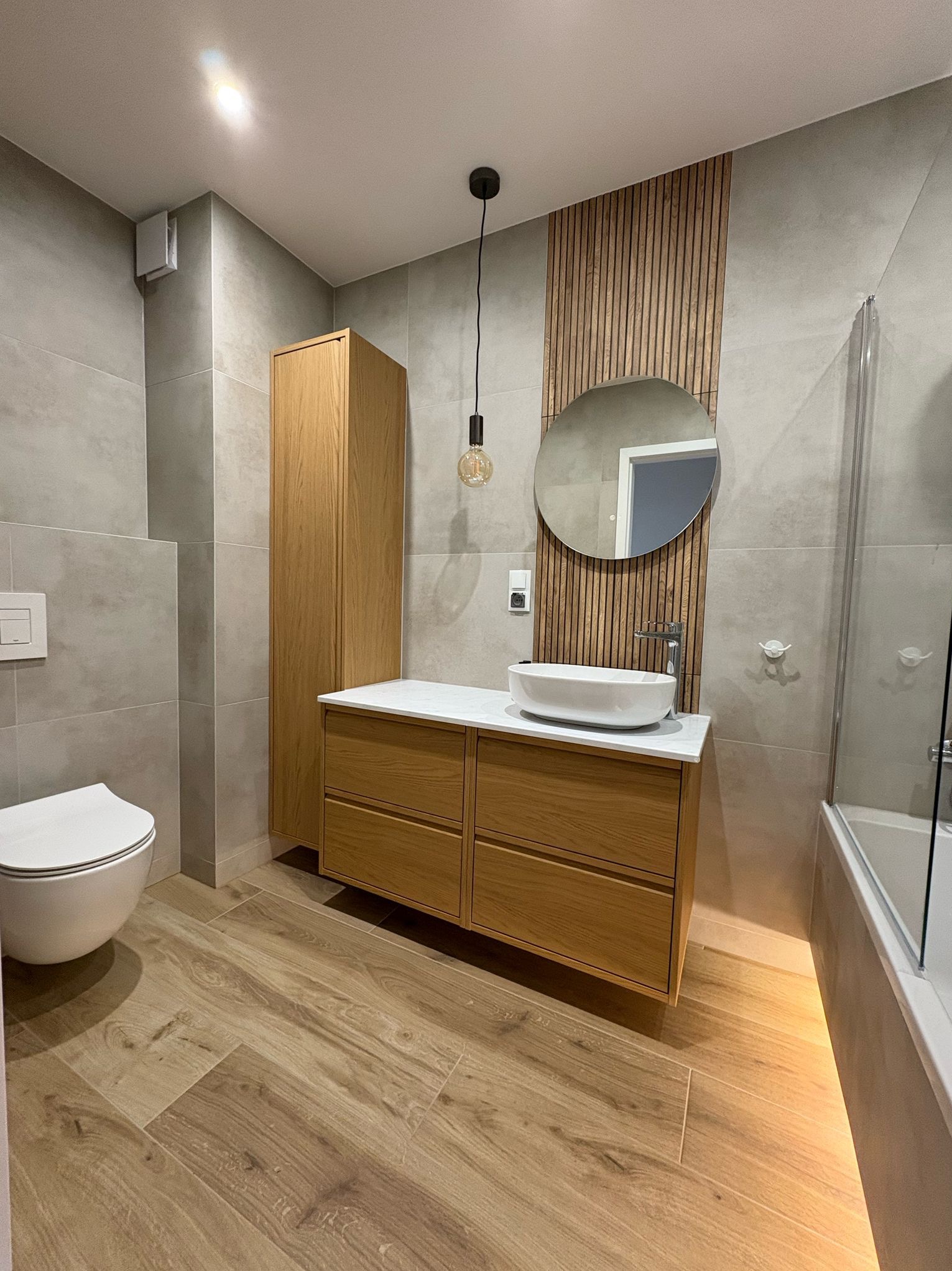 Nowoczesna łazienka z drewnianymi akcentami: szafka pod umywalką z szufladami, wysoka szafka, okrągłe lustro na tle drewnianych paneli, umywalka nablatowa, toaleta podwieszana i podświetlenie LED...