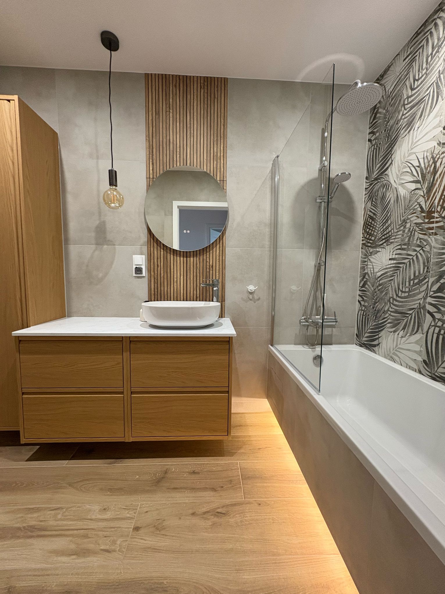 Nowoczesna łazienka z drewnianymi akcentami: szafka pod umywalkę z jasnego drewna, okrągłe lustro, dekoracyjna ściana z drewnianych listew, wanna z parawanem i płytki z motywem liści palmowych.