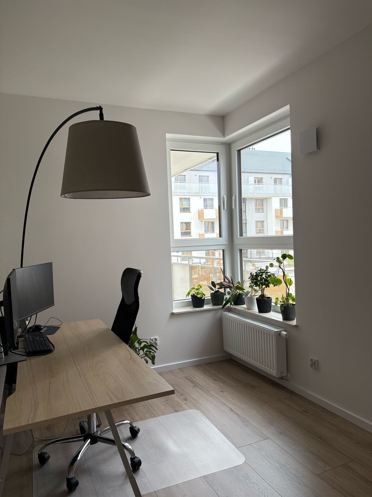 Jasne biuro domowe z drewnianym biurkiem, krzesłem, lampą łukową i parapetem z roślinami doniczkowymi na tle okna z widokiem na budynek mieszkalny.