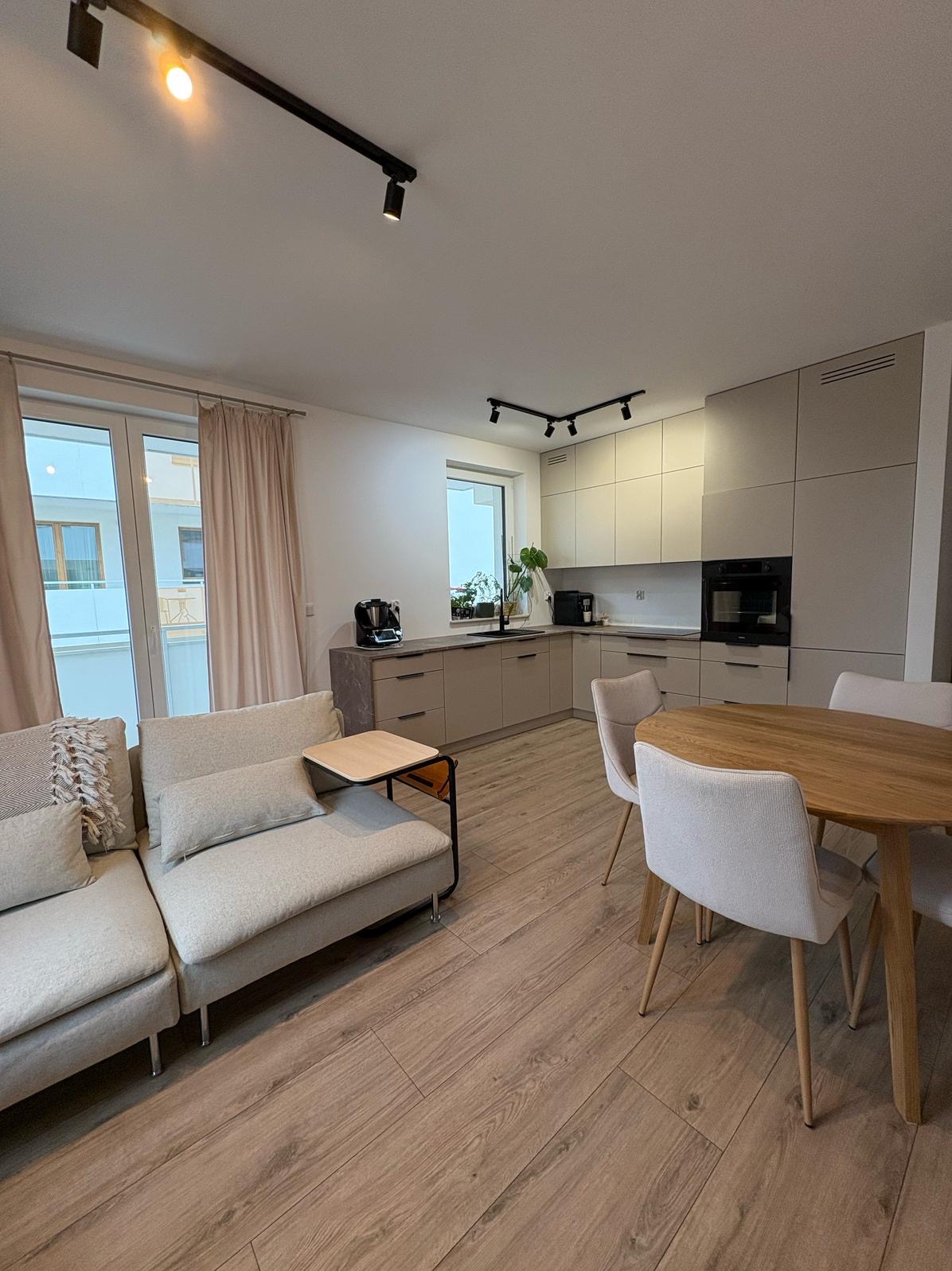 Jasne, nowoczesne wnętrze z drewnianą podłogą, beżową zabudową kuchenną, okrągłym stołem i szarą sofą z małym stolikiem bocznym, oświetlone czarnymi reflektorami sufitowymi.