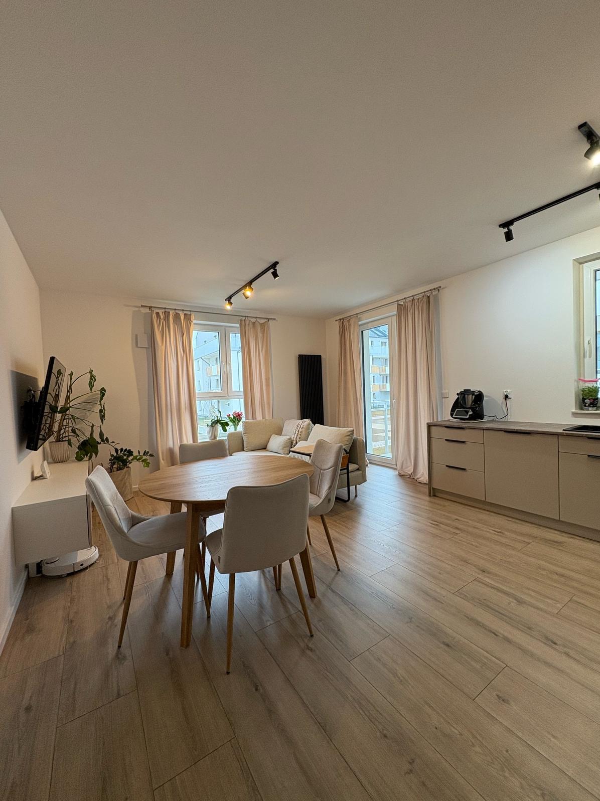 Jasne, przestronne wnętrze mieszkania we Wrocławiu z drewnianą podłogą, okrągłym stołem z krzesłami, beżową sofą i aneksem kuchennym z zabudową w odcieniach szarości, oświetlone reflektorami.