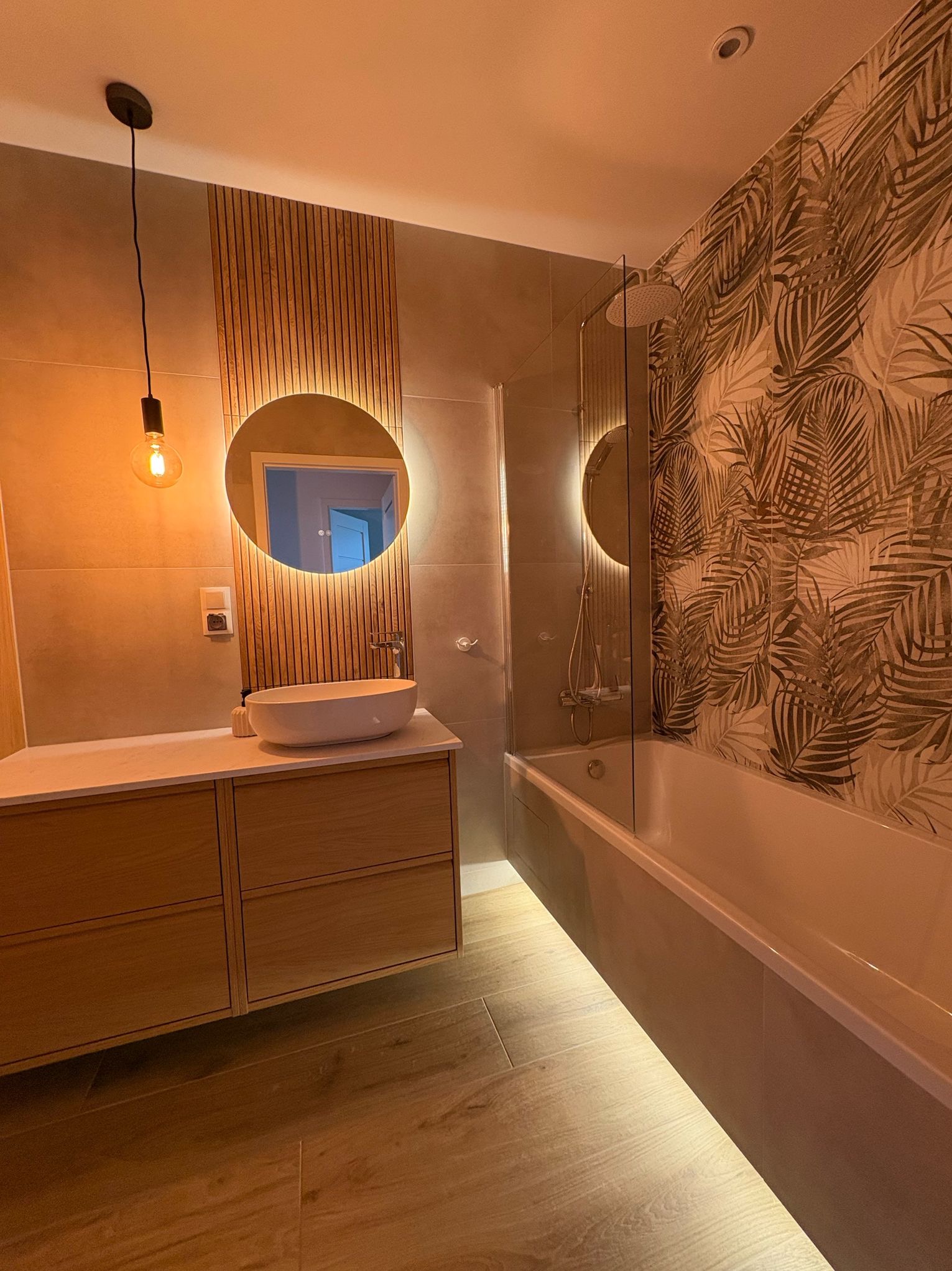 Nowoczesna łazienka z drewnianymi akcentami: okrągłe lustro z podświetleniem LED, umywalka nablatowa, drewniana szafka pod umywalką, wanna z parawanem i tapeta w motywy roślinne.