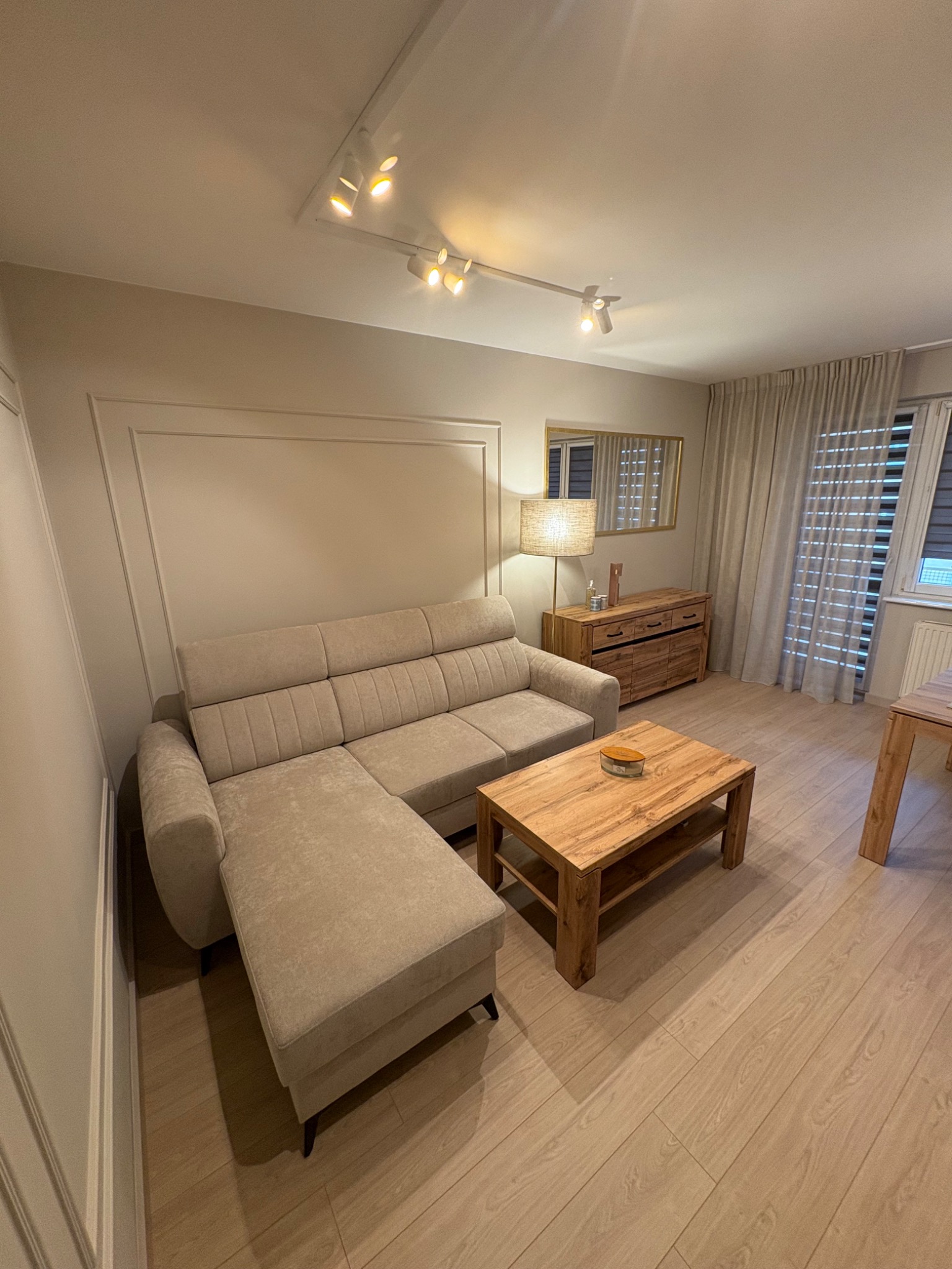 Jasny salon z beżową sofą narożną, drewnianym stolikiem kawowym i komodą. Ściany zdobią listwy dekoracyjne. Parkiet w odcieniach beżu. Oświetlenie punktowe.