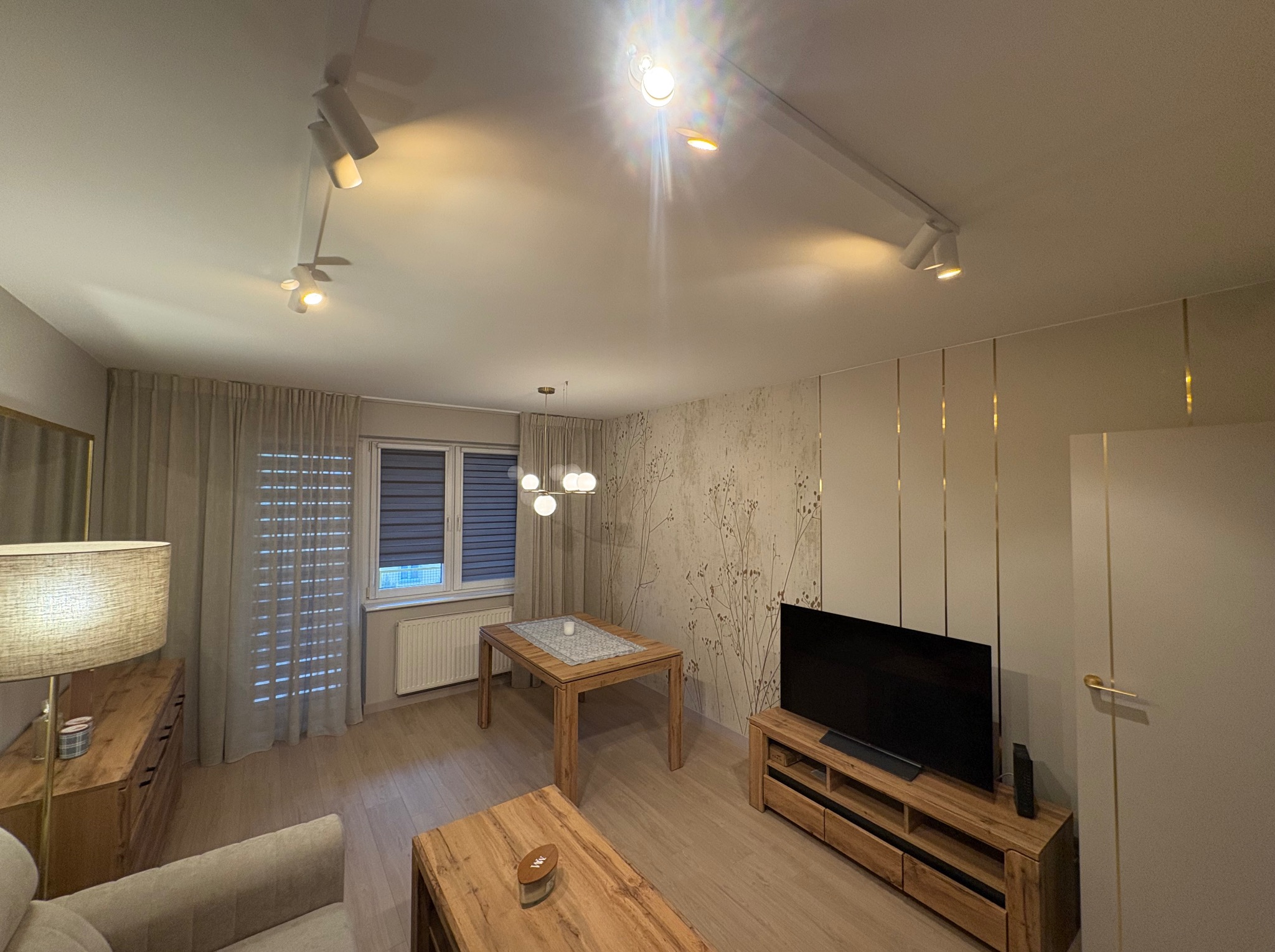 Jasne wnętrze salonu z drewnianymi meblami, oświetleniem punktowym i dekoracyjną tapetą w kwiatowe motywy. Widoczne detale wykończeniowe, takie jak złote listwy.