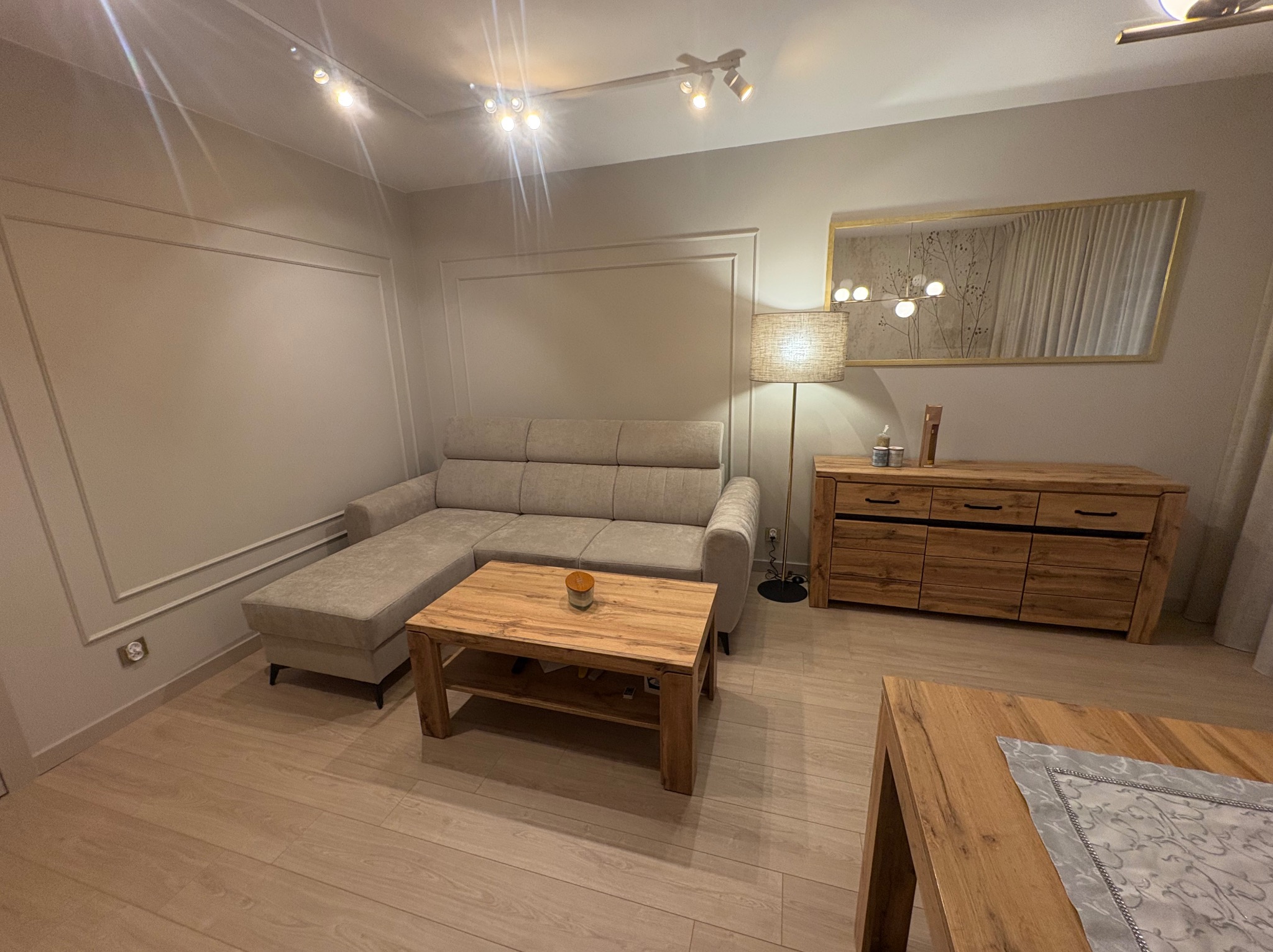 Jasne wnętrze salonu z drewnianymi meblami, beżową sofą i dekoracyjną sztukaterią na ścianach. Parkiet w odcieniach beżu. Lampa podłogowa z abażurem.