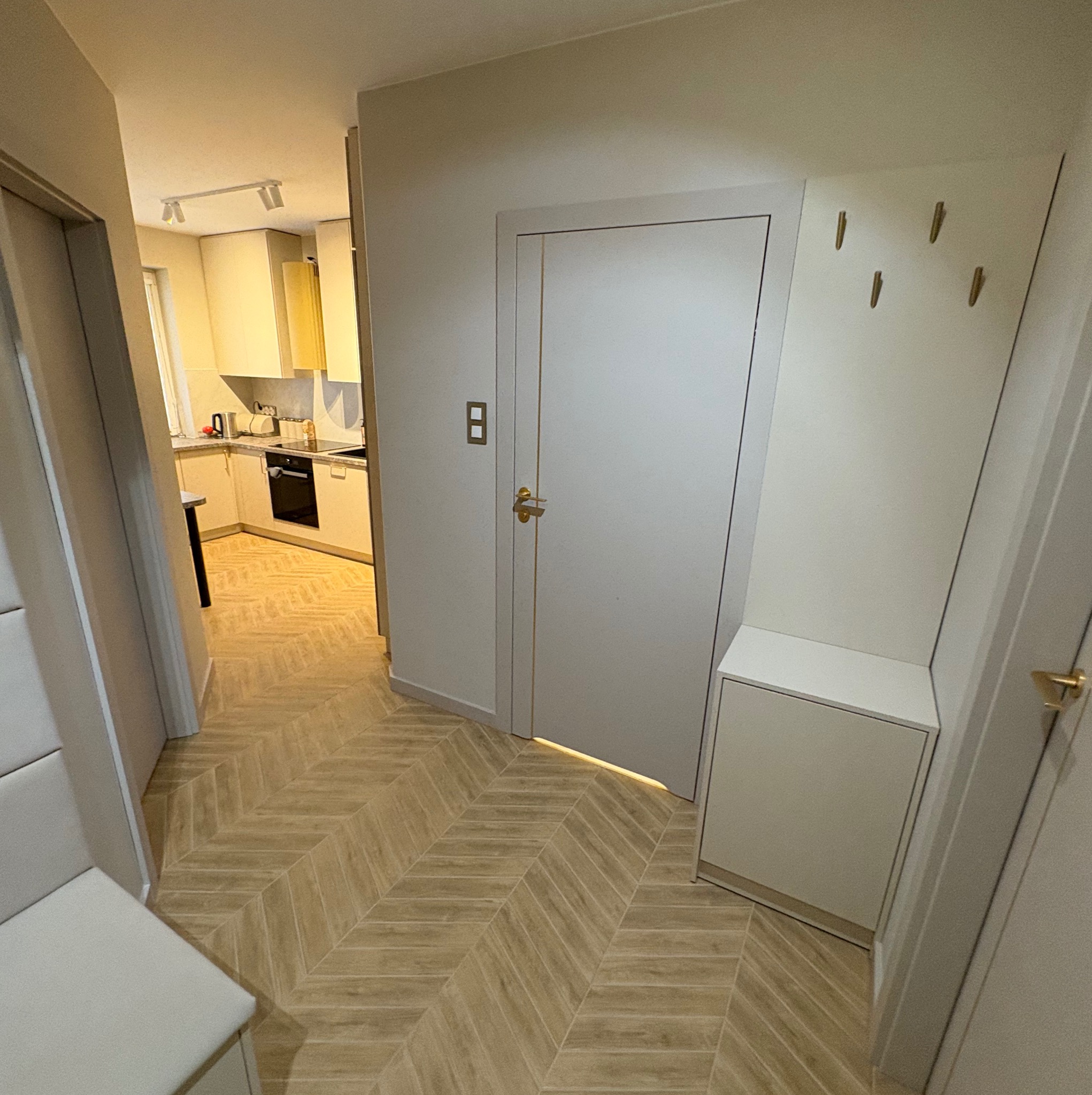 Jasne panele ułożone w jodełkę w korytarzu z widokiem na kuchnię. Minimalistyczne wnętrze z białymi ścianami i drzwiami. Haczyki na ścianie i szafka na buty.