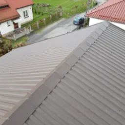 Nowy, brązowy dach z blachy trapezowej, widok z góry, w tle domy i droga z zaparkowanym niebieskim samochodem.