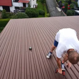 Pracownik w białej koszulce i trampkach montuje brązowe blaszane pokrycie dachowe za pomocą wkrętarki, w tle domy i ulica.