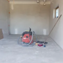 Wnętrze w trakcie remontu, widoczny czerwony odkurzacz przemysłowy, ściany po szpachlowaniu, podłoga z resztkami gładzi, okno z butelką wody, zakryte folią fragmenty ścian.
