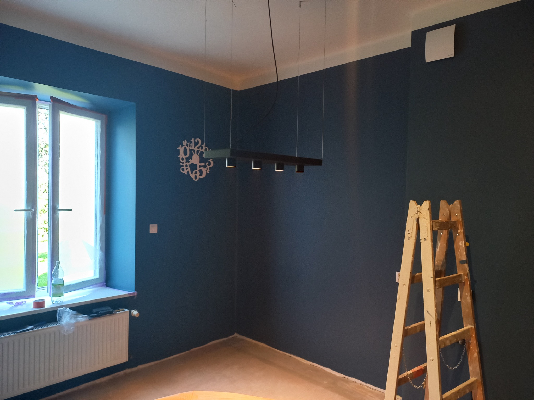 Pokój po malowaniu ścian na ciemny niebieski kolor, z drewnianą drabiną opartą o ścianę, nowoczesną lampą wiszącą i ozdobnym zegarem na ścianie, widoczne zabezpieczenia okna folią malarską.