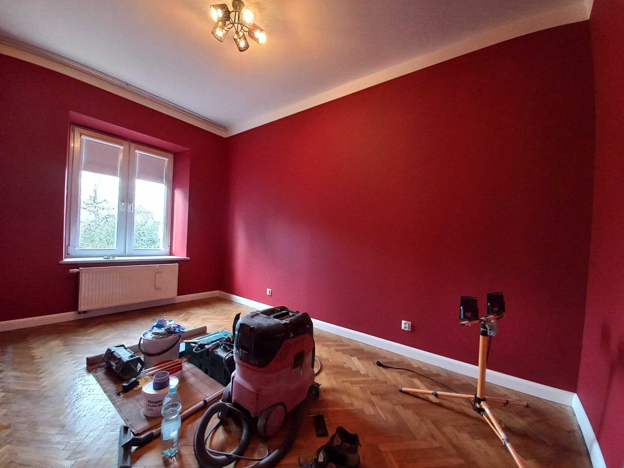 Pokój po malowaniu ścian na intensywny czerwony kolor, widoczne narzędzia malarskie, lampa budowlana i odkurzacz przemysłowy na parkiecie, okno z roletami w tle.