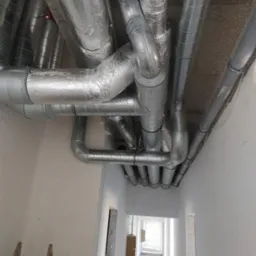 Widok z dołu na system rur wentylacyjnych pod sufitem w surowym wnętrzu, część z izolacją aluminiową, w tle widoczny korytarz z otwartymi drzwiami i drabina oparta o ścianę.