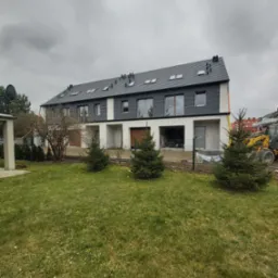 Trzy segmenty szeregowe w trakcie budowy, z szarą elewacją i ciemnym dachem, widoczne okna dachowe, teren wokół częściowo zagospodarowany, na trawniku trzy małe świerki, w tle żółta koparka.