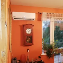 Biała klimatyzacja marki Kaisai zamontowana na pomarańczowej ścianie w pokoju, obok wisi drewniany zegar z wahadłem, a pod spodem stoi czarny stolik z dekoracjami. W tle widoczne okno z firanką...