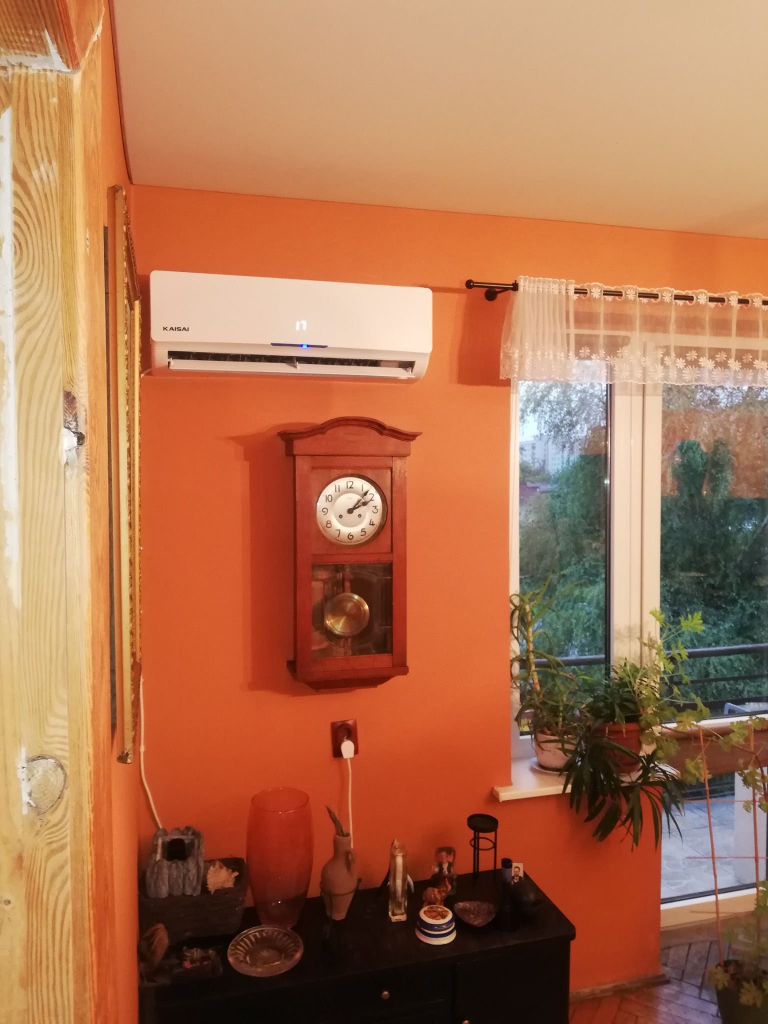 Biała klimatyzacja marki Kaisai zamontowana na pomarańczowej ścianie w pokoju, obok wisi drewniany zegar z wahadłem, a pod spodem stoi czarny stolik z dekoracjami. W tle widoczne okno z firanką...