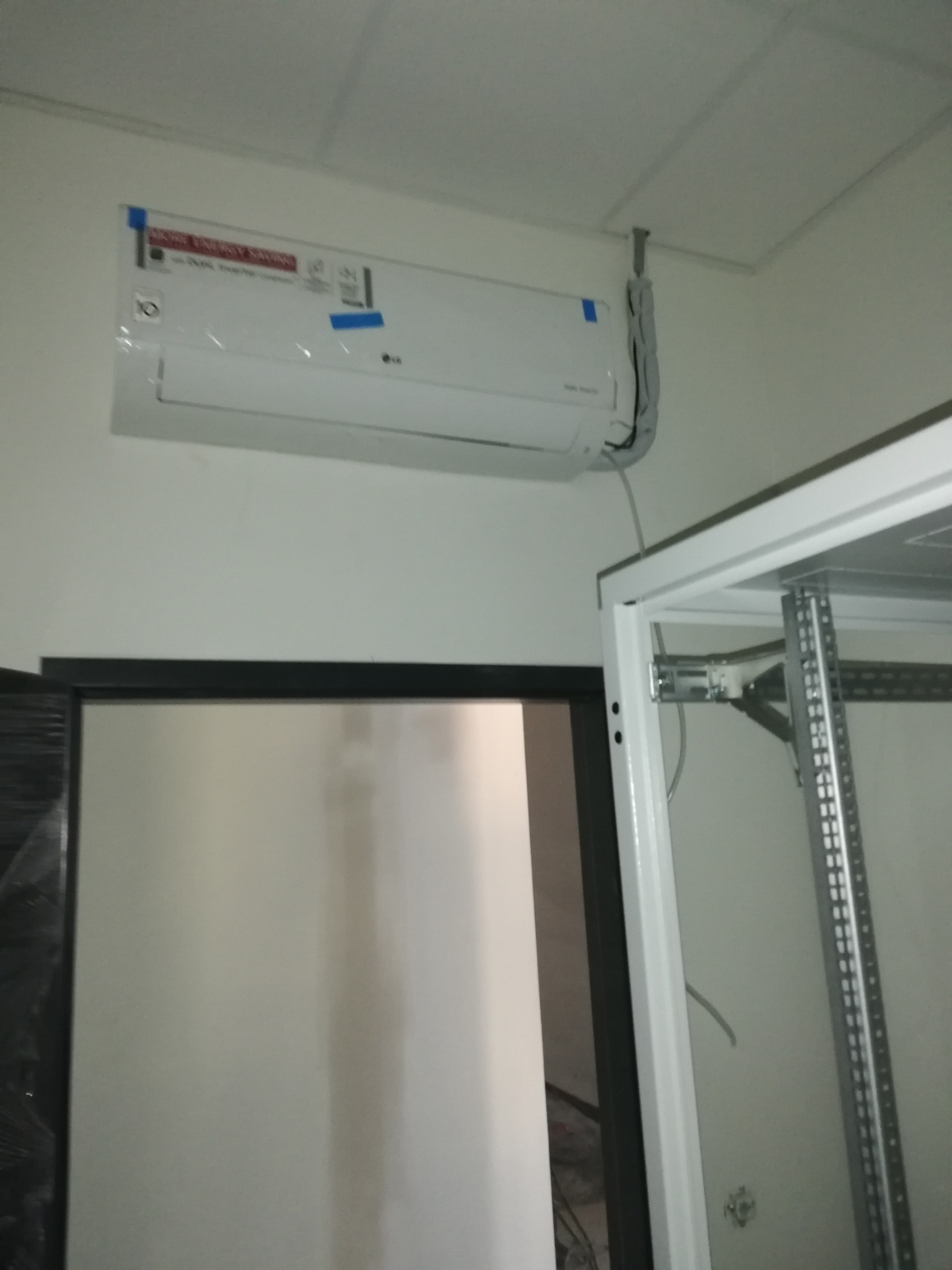 Biała jednostka wewnętrzna klimatyzacji LG zamontowana na ścianie, z widocznymi niebieskimi taśmami zabezpieczającymi i przewodami w szarej osłonie, obok metalowej konstrukcji regału.