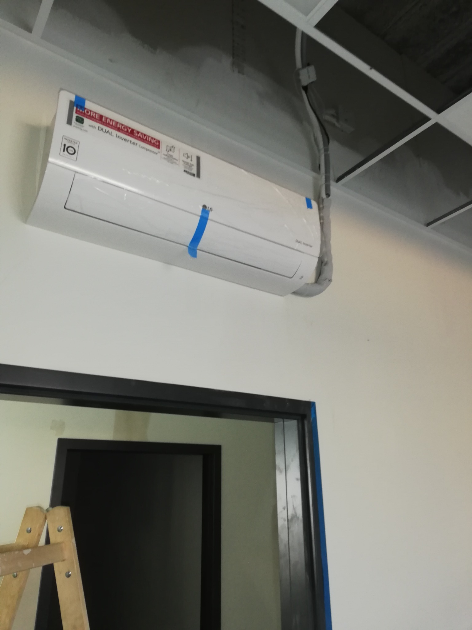 Biała klimatyzacja LG z niebieską taśmą montażową, widoczne przewody i drabina malarska w pomieszczeniu biurowym, instalacja w trakcie.