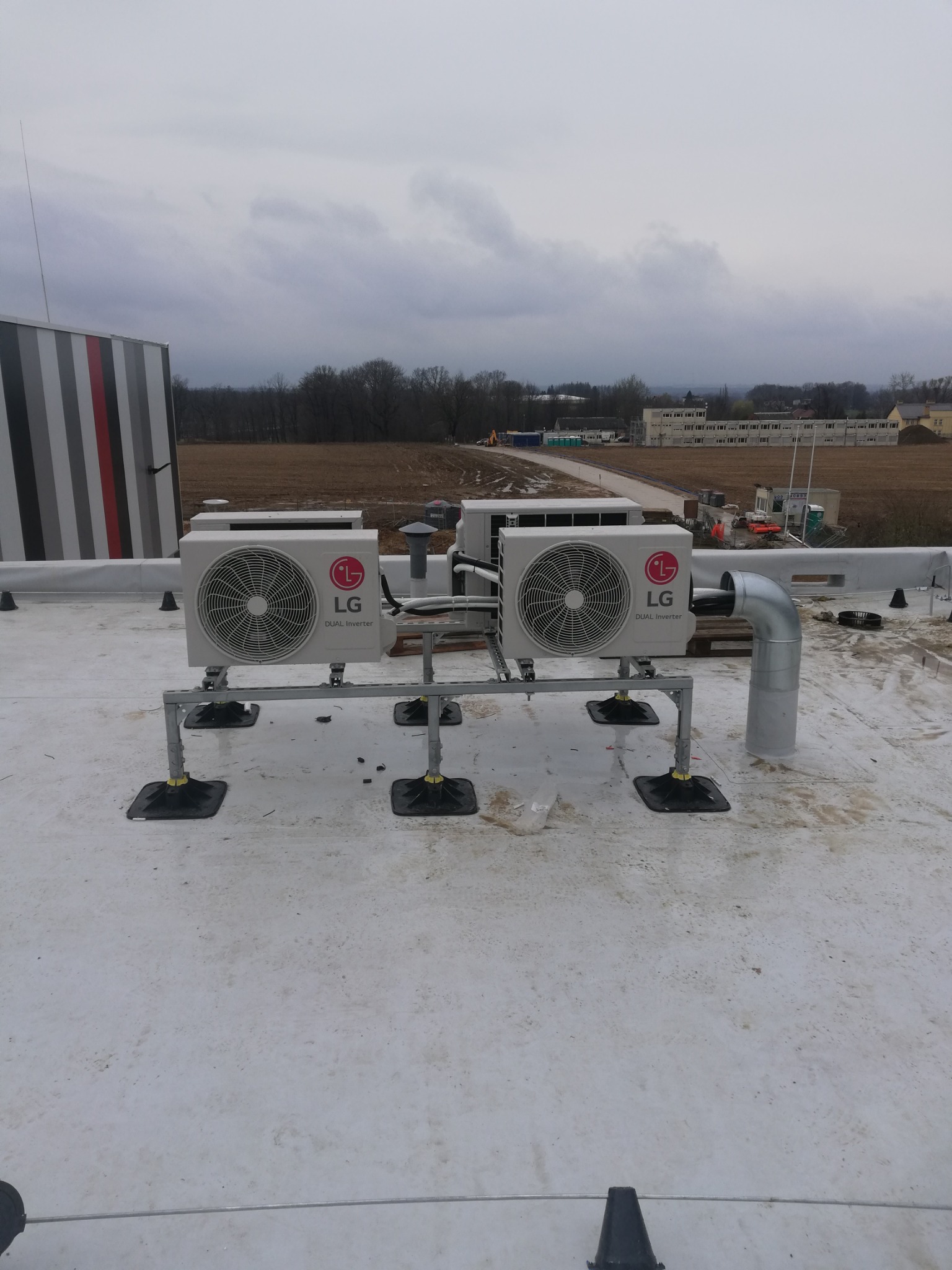 Dwa agregaty klimatyzacyjne LG Dual Inverter, zamontowane na metalowych konstrukcjach z czarnymi podstawami, na płaskim dachu budynku komercyjnego, z widocznymi elementami instalacji wentylacyjnej...