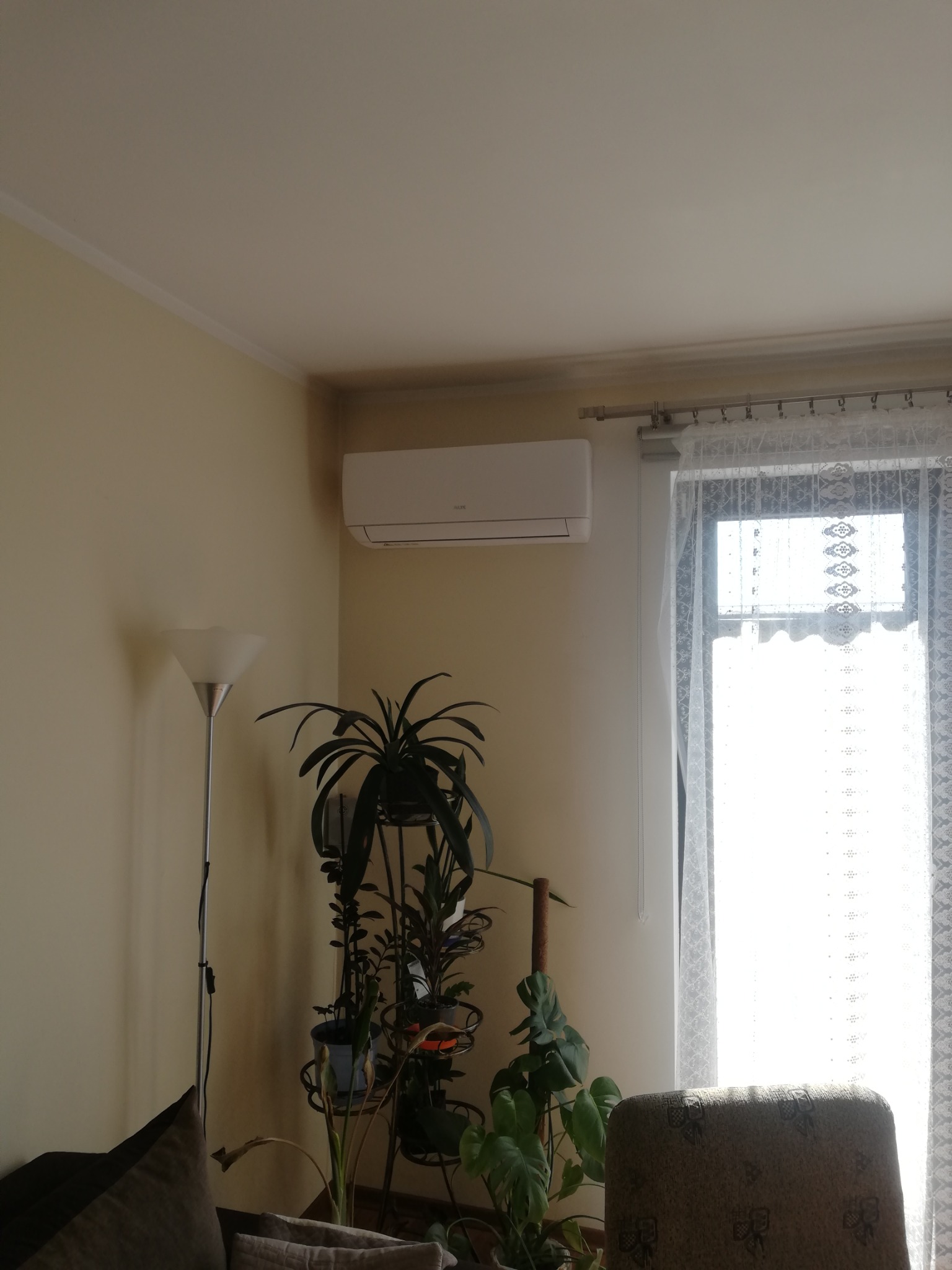 Biały klimatyzator ścienny marki AUX zamontowany w rogu pokoju nad zielonymi roślinami w doniczkach, obok okna z firanką.