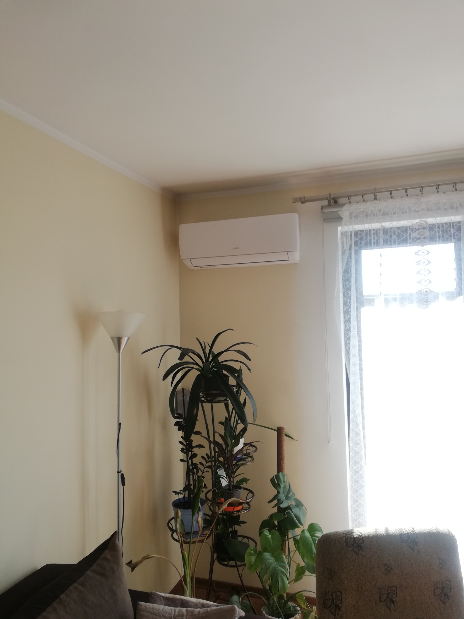 Biały klimatyzator ścienny zamontowany w jasnym pokoju, obok okna z firanką i wysokiej lampy podłogowej, z roślinami doniczkowymi na metalowym kwietniku.