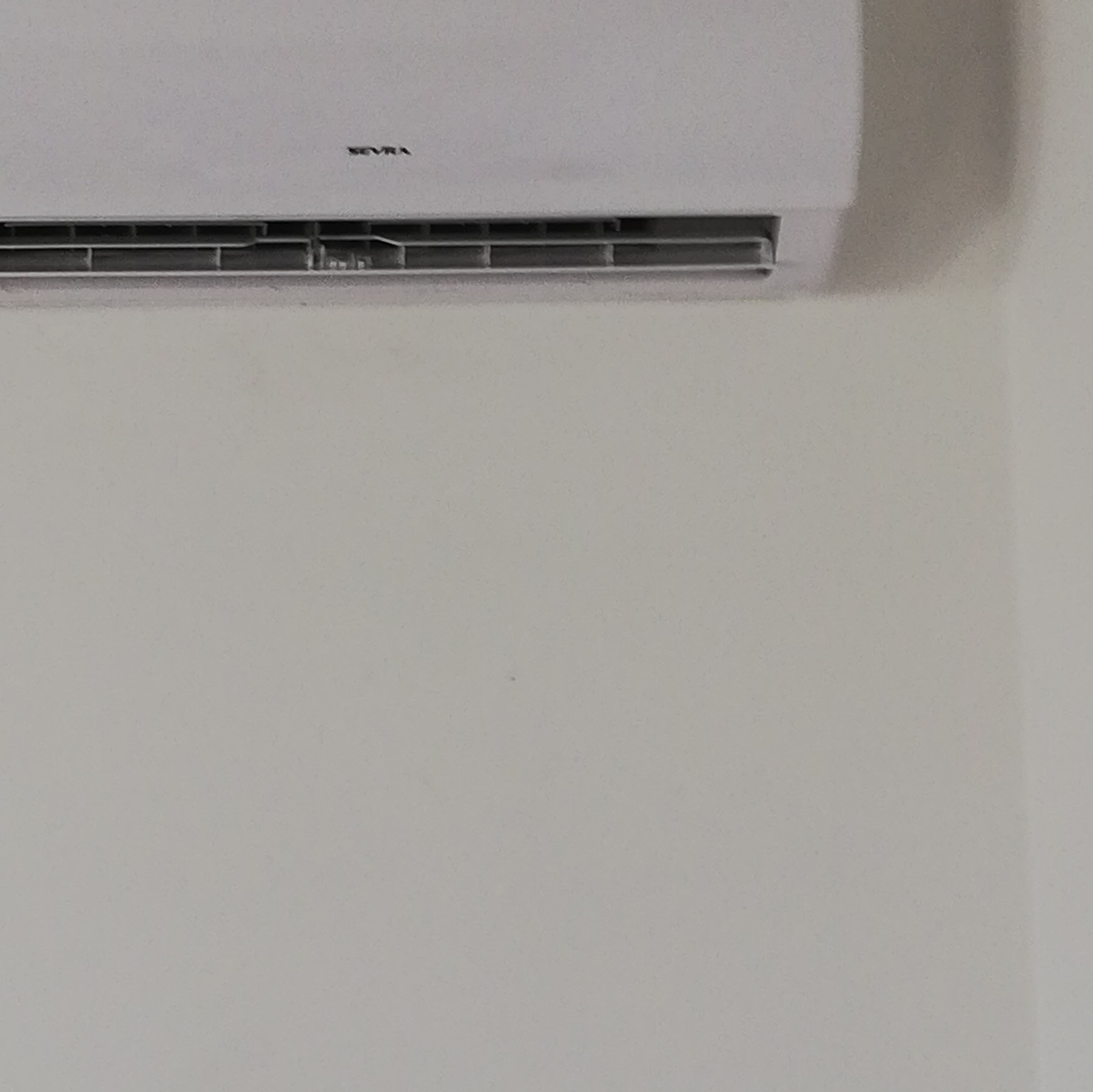 Biały klimatyzator marki SEVRA zamontowany na jasnej ścianie, widok z bliska na górną część urządzenia.