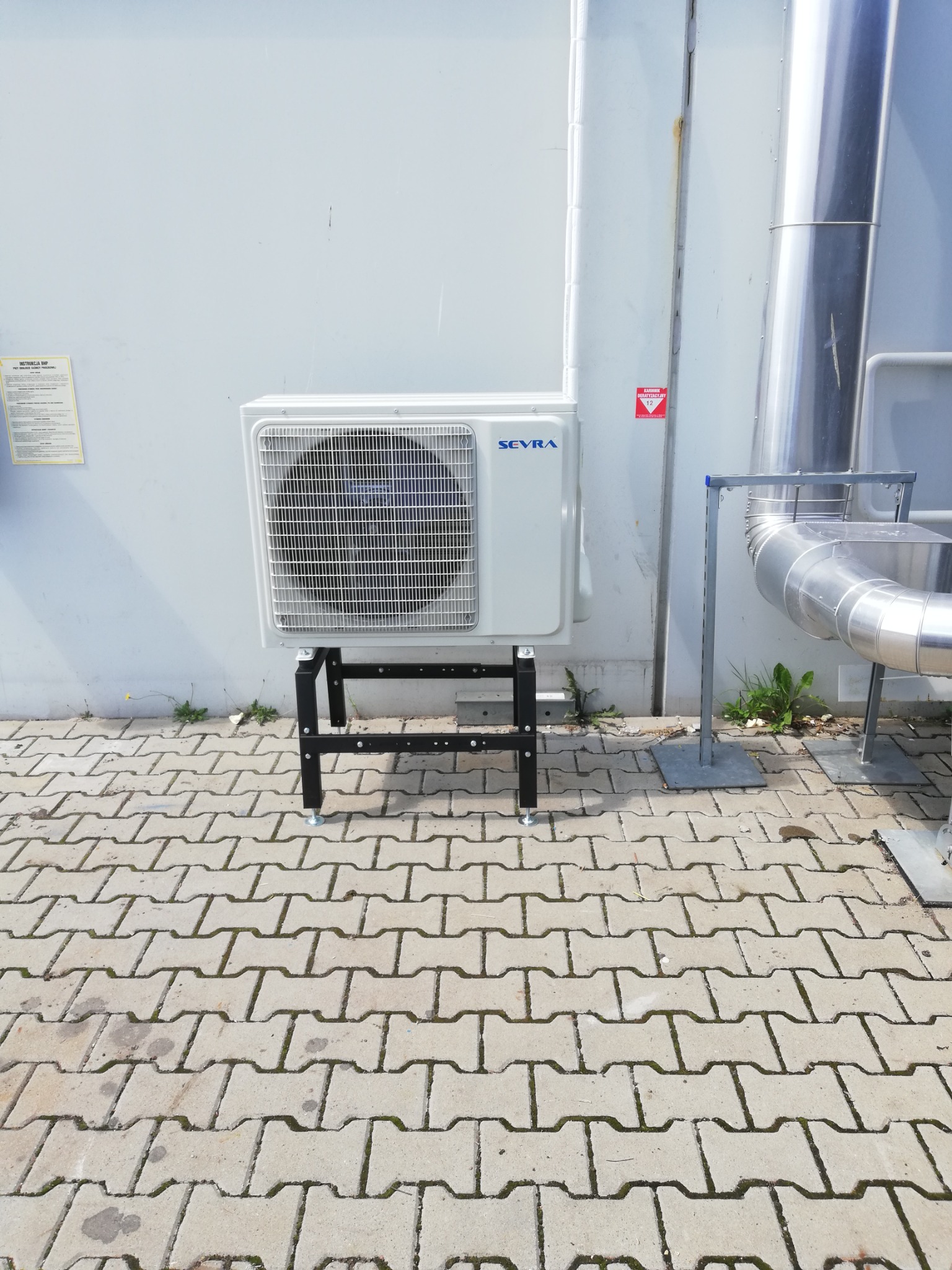 Zewnętrzna jednostka klimatyzacji marki Sevra, zamontowana na czarnym, metalowym stelażu, stoi na kostce brukowej przy jasnej ścianie budynku. Obok widoczne rury wentylacyjne.