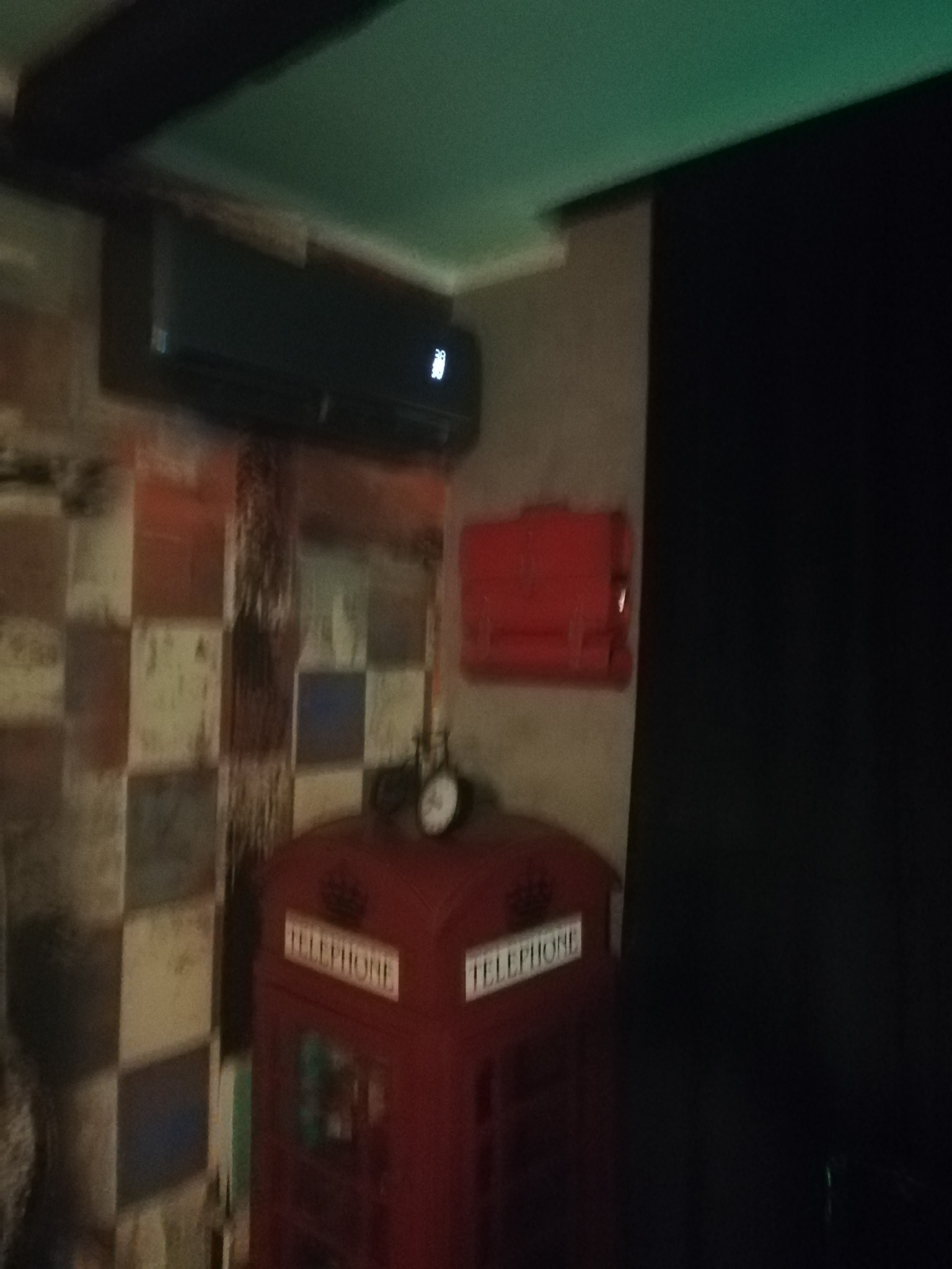 Czarna jednostka klimatyzacyjna zamontowana na ścianie nad dekoracyjną czerwoną budką telefoniczną, widoczna na tle ściany w kratkę.