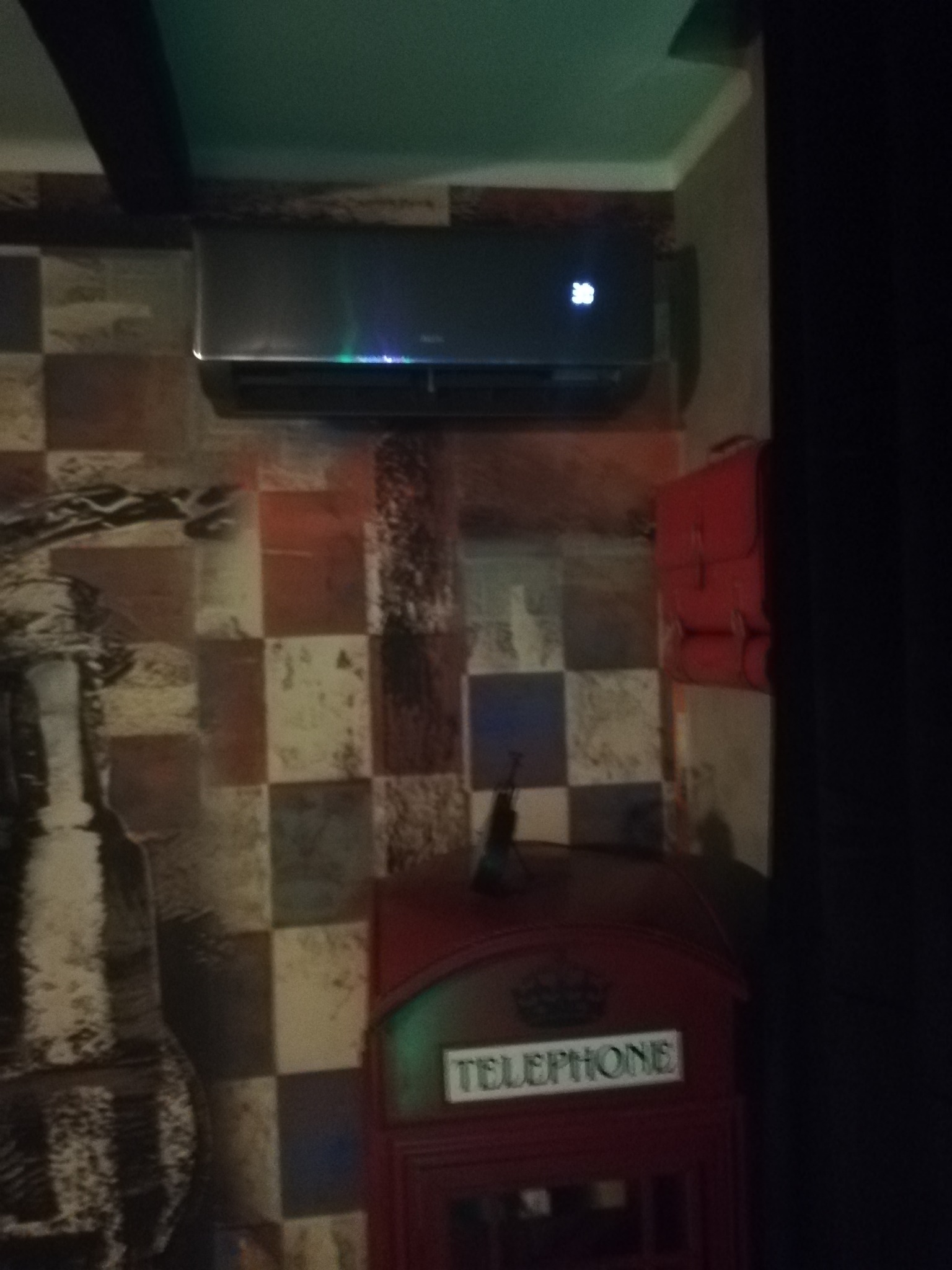 Czarny klimatyzator ścienny z wyświetlaczem temperatury 20 stopni, zainstalowany na ścianie z motywem szachownicy, obok czerwonej budki telefonicznej i czerwonej torby.