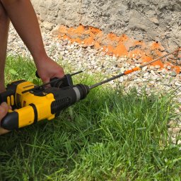 T&K BUDOWNICTWO KAMIL KOSZYKOWSKI - Praca z wiertarką udarową przy fundamencie, widoczna linia izolacyjna i trawa. Detal narzędzia i fragmentu ściany.