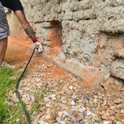 T&K BUDOWNICTWO KAMIL KOSZYKOWSKI - Iniekcja krystaliczna w ścianę z cegły i betonu. Pracownik w rękawicach wprowadza preparat przez wężyk do otworów w murze. Kamieniste podłoże.
