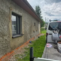 Elewacja budynku z szorstkim tynkiem, widoczne narzędzia i kompresor na przyczepie. Przygotowania do prac tynkarskich, ceglana podmur&oacute;wka.