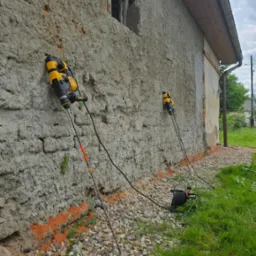 Iniekcja krystaliczna w starym, ceglanym budynku. Dwa urządzenia iniekcyjne przymocowane do ściany, z widoczną perforacją i przewodami. Otoczenie: kamienie i trawa.
