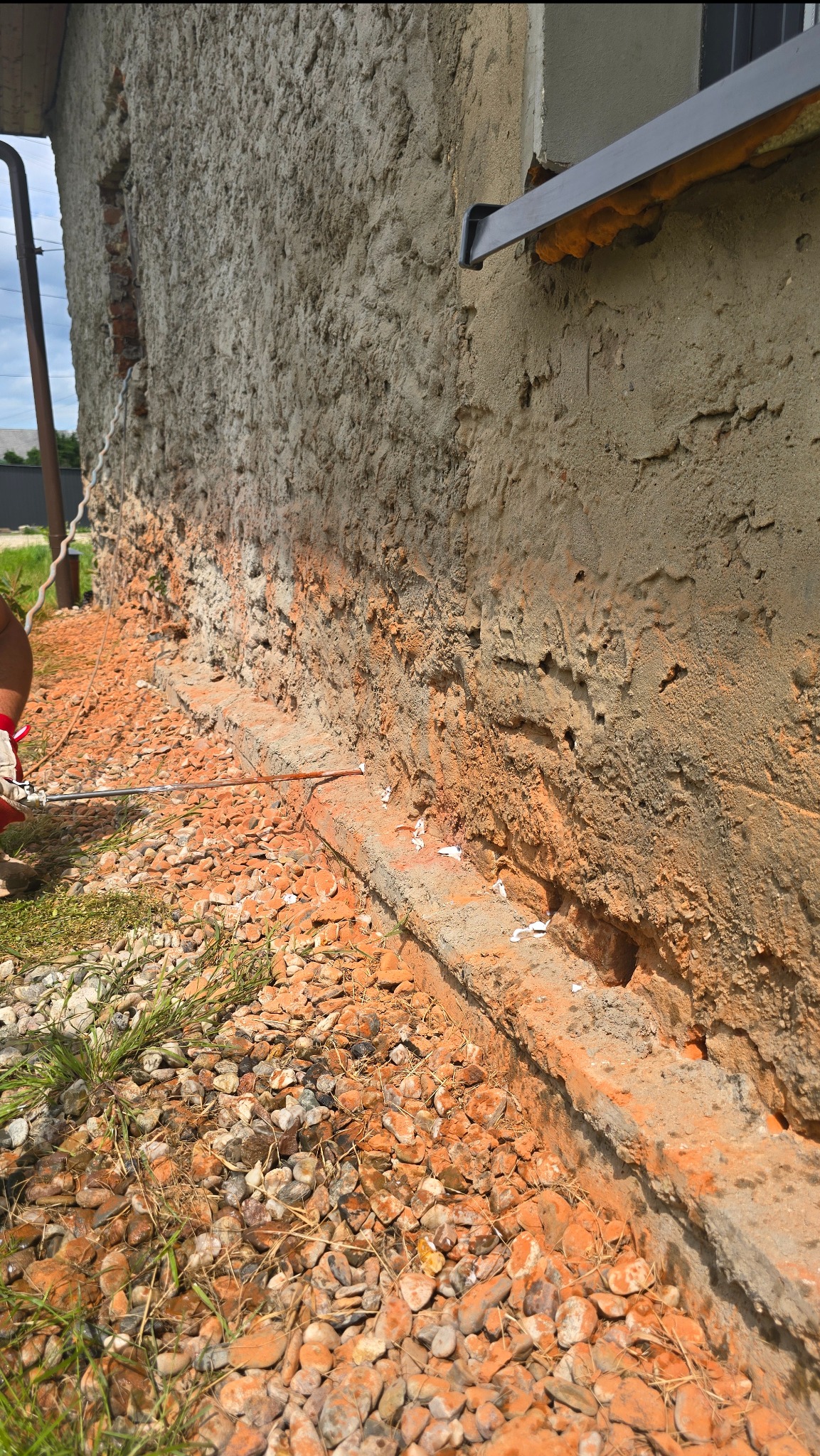 Iniekcja krystaliczna w murze fundamentowym, widoczne nawiercone otwory i aplikator. Kamienie ozdobne wokół fundamentu. Fragment ściany pokryty tynkiem.