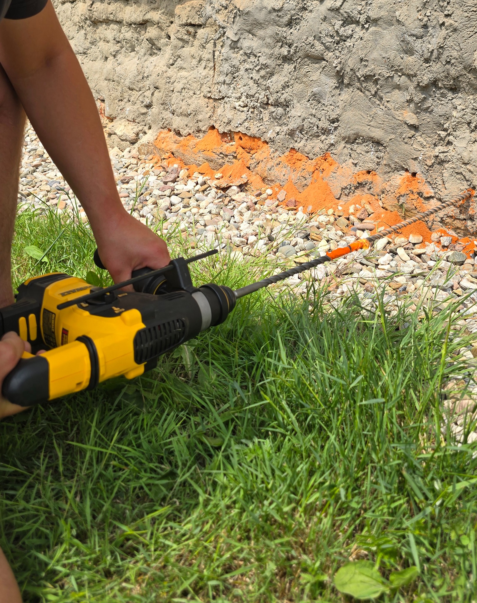 Praca z wiertarką udarową przy fundamencie, widoczna linia izolacyjna i trawa. Detal narzędzia i fragmentu ściany.