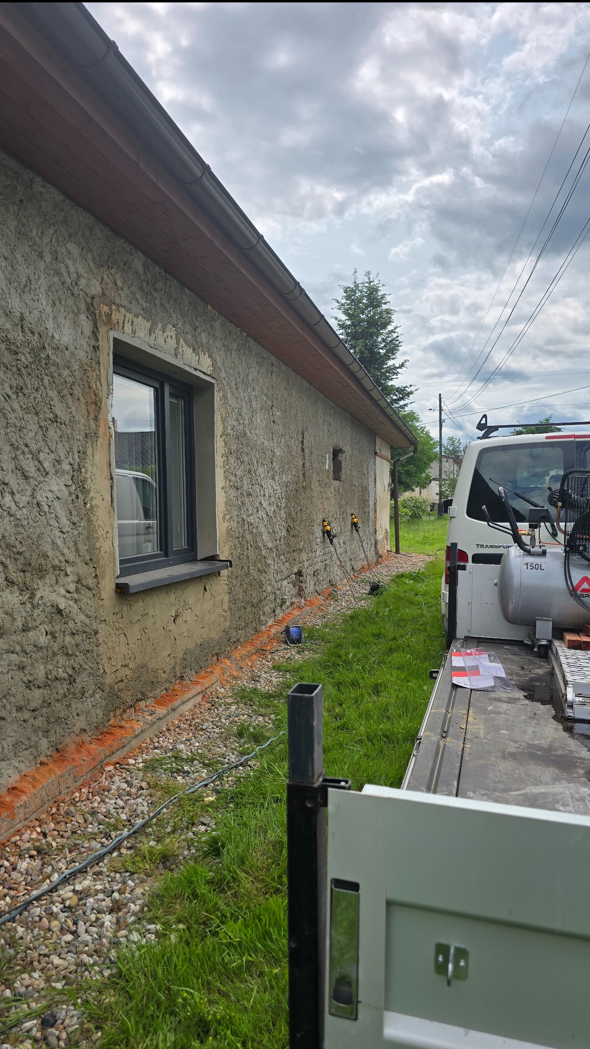 Elewacja budynku z szorstkim tynkiem, widoczne narzędzia i kompresor na przyczepie. Przygotowania do prac tynkarskich, ceglana podmurówka.