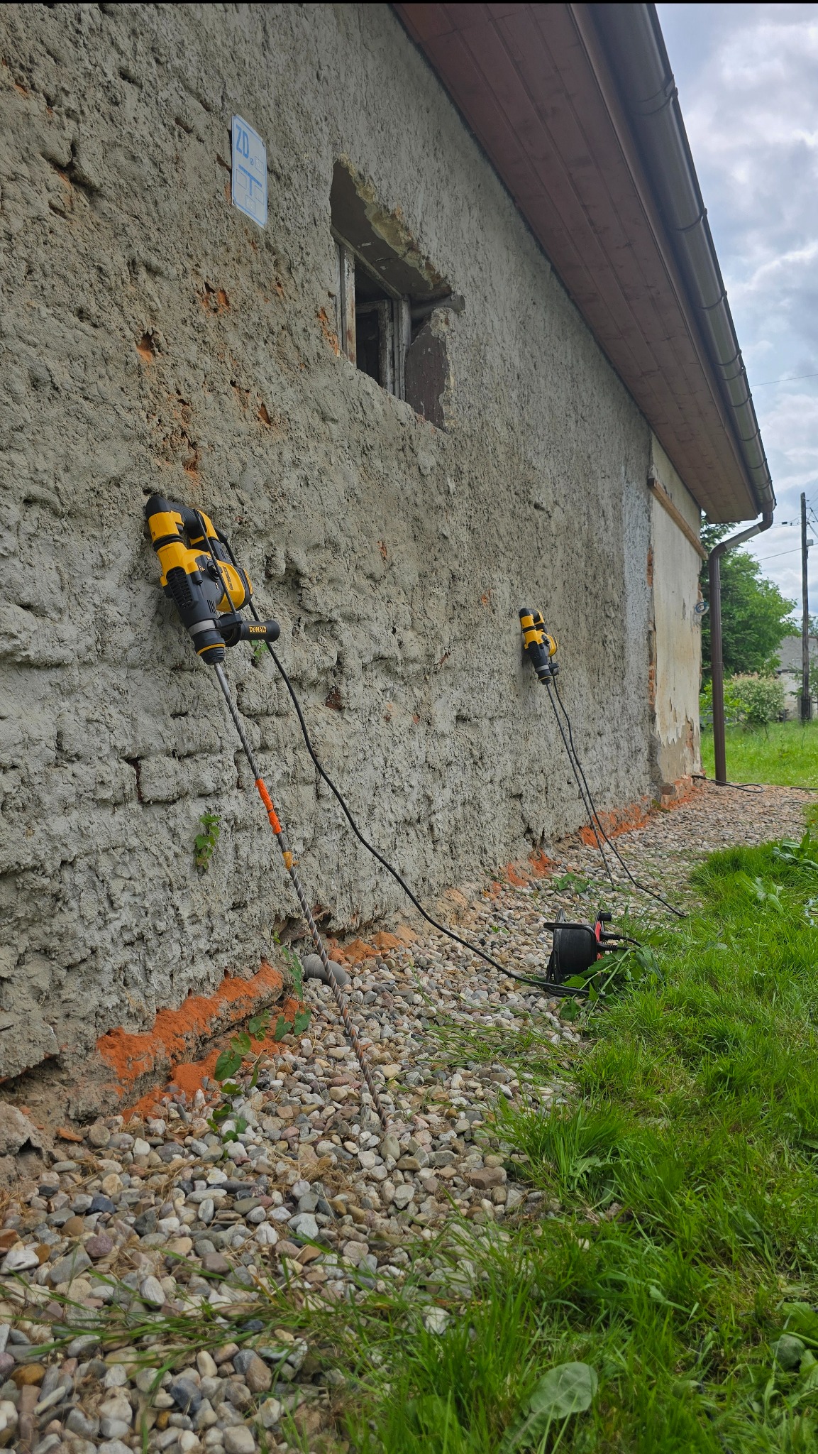Iniekcja krystaliczna w starym, ceglanym budynku. Dwa urządzenia iniekcyjne przymocowane do ściany, z widoczną perforacją i przewodami. Otoczenie: kamienie i trawa.