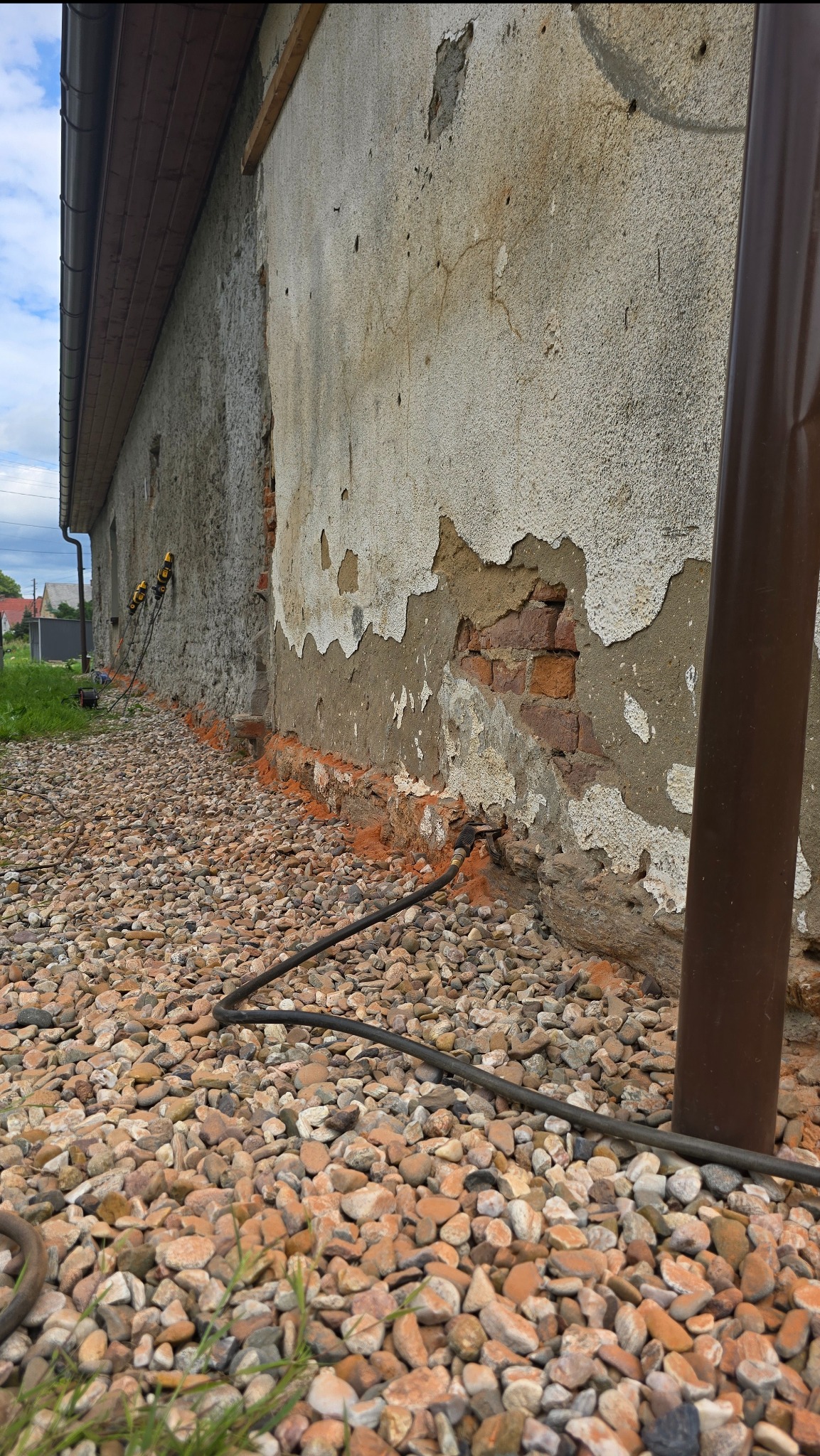 Ściana budynku z uszkodzonym tynkiem, odsłoniętą cegłą i śladami wilgoci. Widoczne narzędzia i kable sugerują prace osuszające.