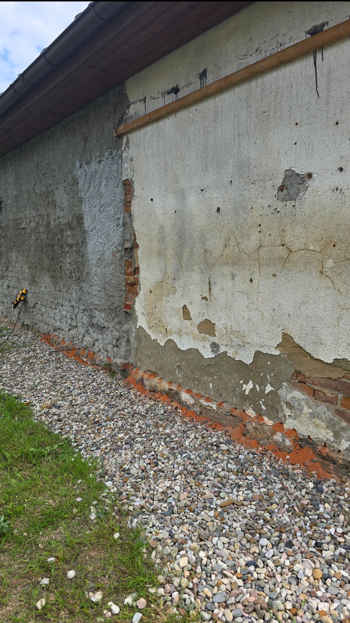 Uszkodzona elewacja budynku z odpadającym tynkiem, odsłaniająca cegły. Widoczne ślady wilgoci i uszkodzeń, kamienny drenaż przy podstawie ściany.