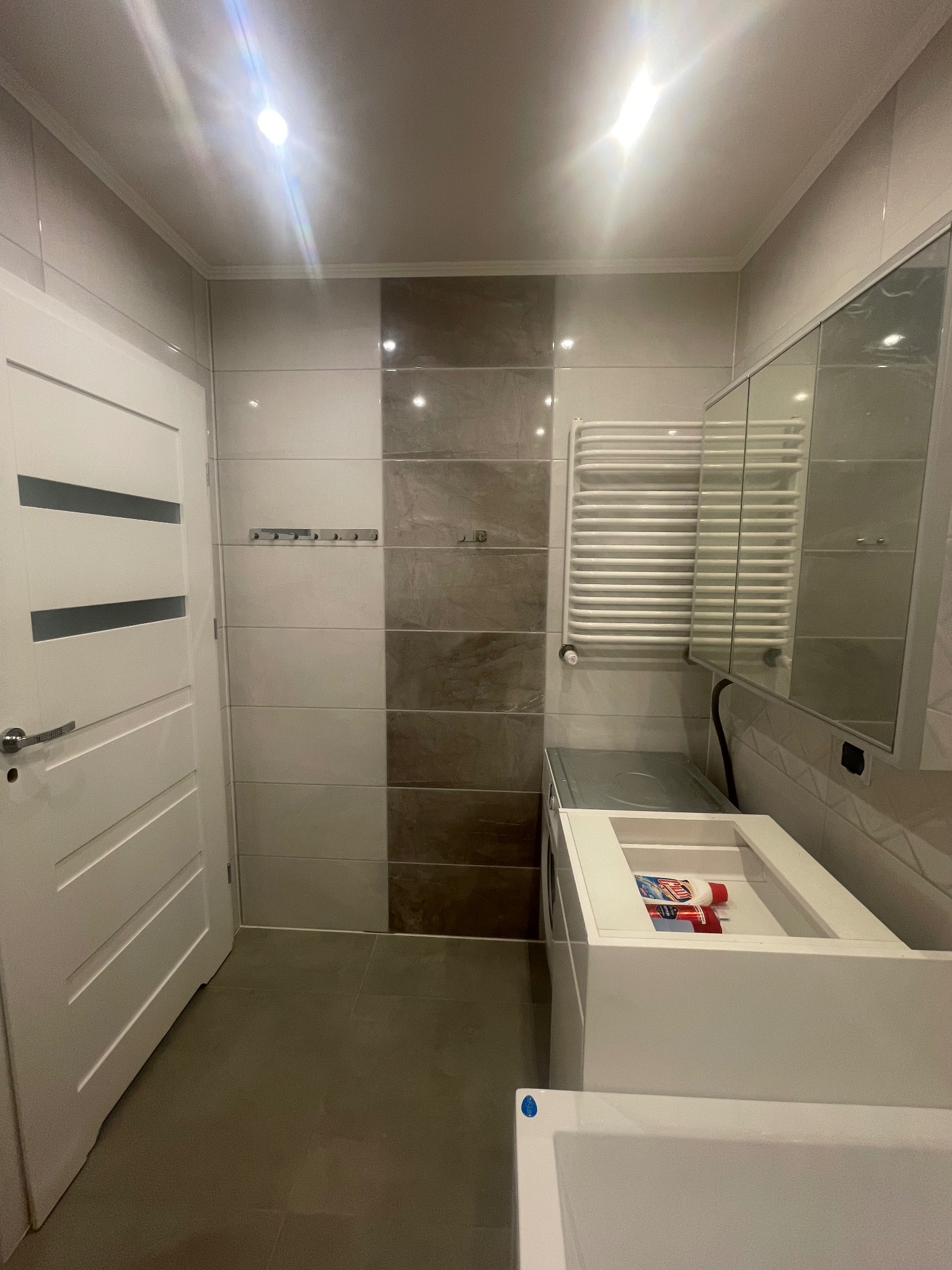 Nowoczesna łazienka z białymi płytkami i akcentem w odcieniach szarości, grzejnik drabinkowy, szafka z umywalką i widoczną chemią gospodarczą, drzwi z poziomymi frezami.