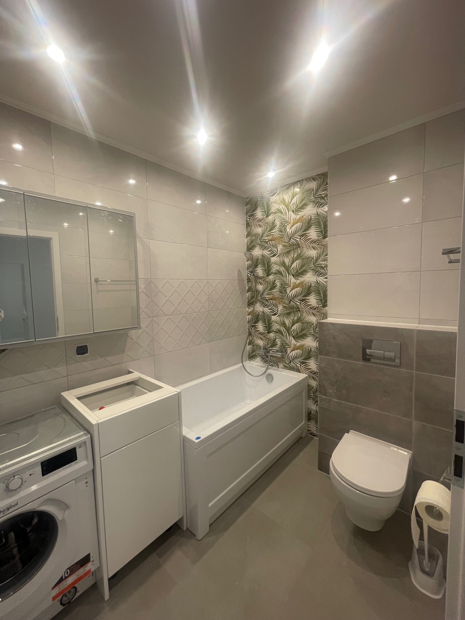 Nowoczesna łazienka z białymi płytkami, akcentem w postaci tapety z motywem liści, białą wanną, toaletą i pralką Whirlpool. Widoczna szafka pod umywalkę bez blatu i lustro z szafką.