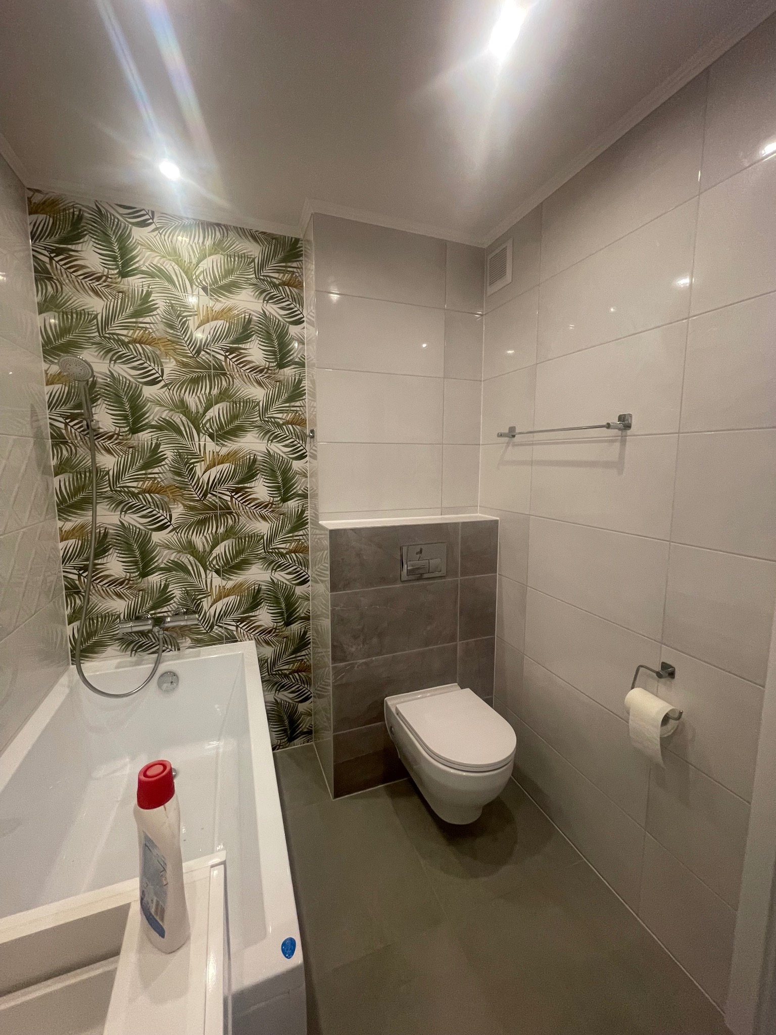 Nowoczesna łazienka z białą armaturą, płytkami w odcieniach szarości i akcentem tropikalnej tapety za wanną, na której stoi butelka środka czyszczącego.