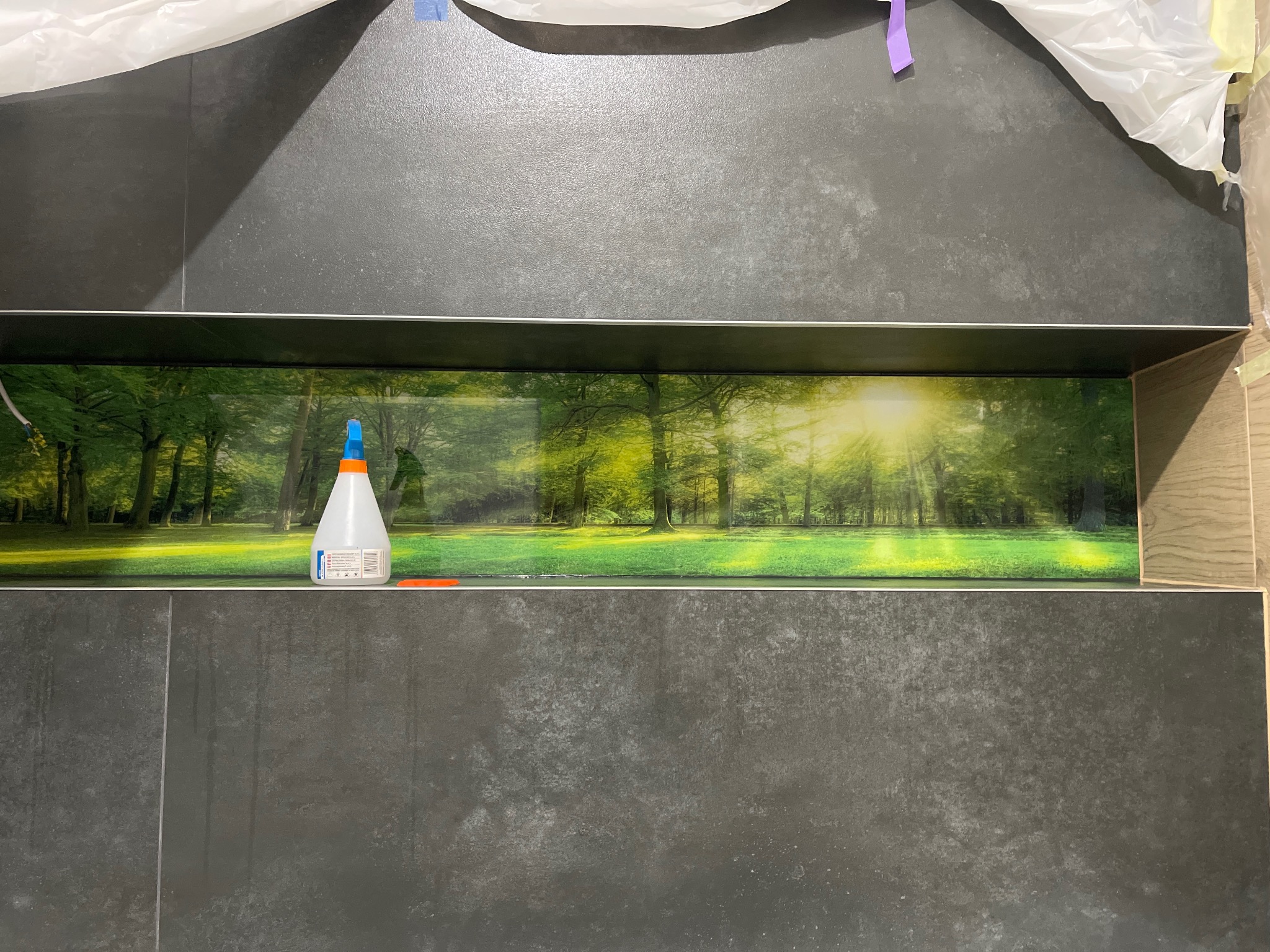 Montaż panelu dekoracyjnego z motywem leśnym na ciemnych płytkach ściennych, widoczna butelka z klejem i fragment folii ochronnej.