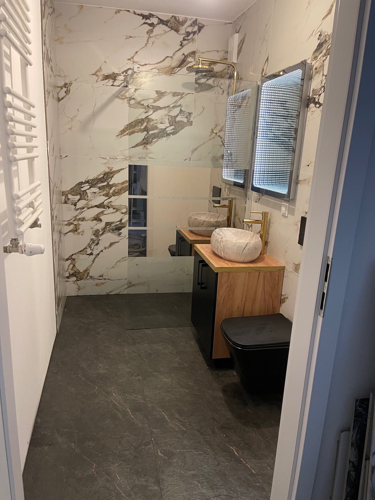 Nowoczesna łazienka z marmurowymi płytkami na ścianach, umywalkami w kształcie kamieni, podświetlanym lustrem i czarną toaletą.