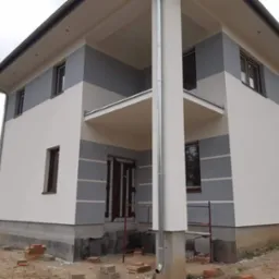 Dwukondygnacyjny dom murowany z ocieploną elewacją w odcieniach bieli i szarości, z widocznymi rusztowaniami i niedokończonym terenem wokół.