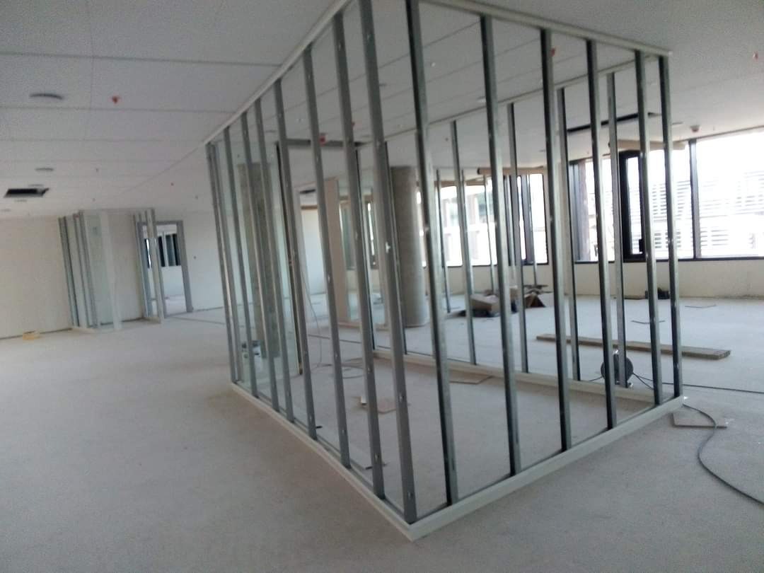 Metalowa konstrukcja ścian działowych w surowym stanie w przestronnym, jasnym pomieszczeniu biurowym, widoczne okna i podłoga w trakcie prac wykończeniowych.