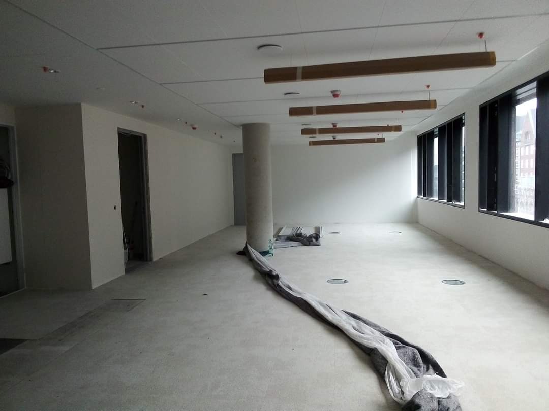 Duże, puste pomieszczenie biurowe w trakcie prac wykończeniowych; widoczne surowe ściany, betonowa podłoga, kolumna i prowizoryczne oświetlenie sufitowe, okna z czarnymi ramami oraz zabezpieczenia...