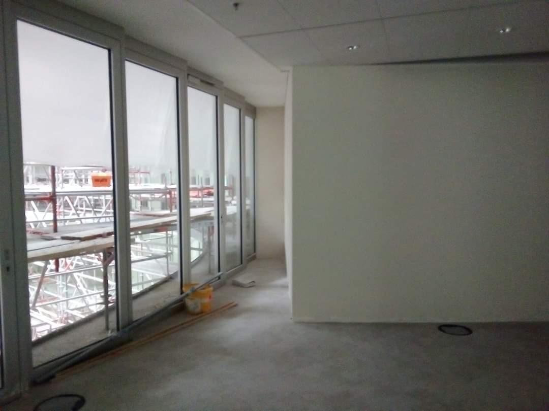 Wnętrze w stanie deweloperskim z widokiem na rusztowania przez duże okna przesuwne, widoczne rury i wiadro na podłodze.